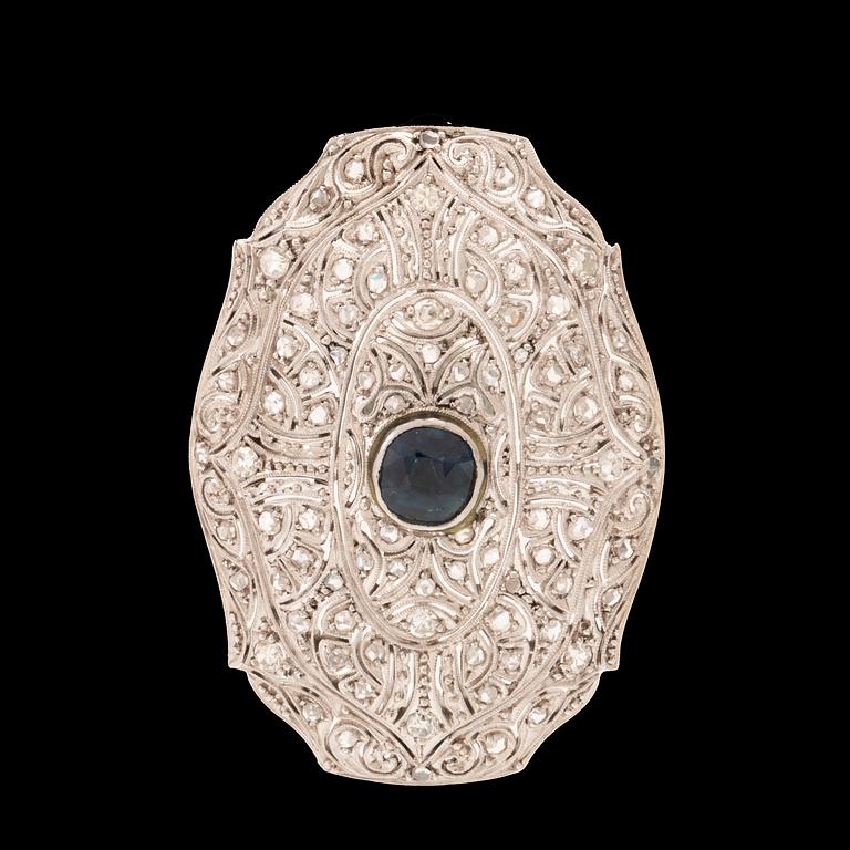 An 18K white gold brooch set with a facet-cut sapphire possibly synthetic, as well as brilliant and rose-cut diamonds.