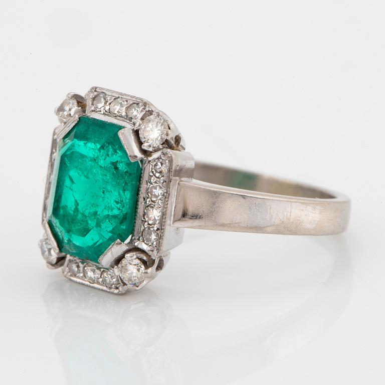 An 18K white gold ring set with a faceted emerald and round brilliant-cut diamonds.