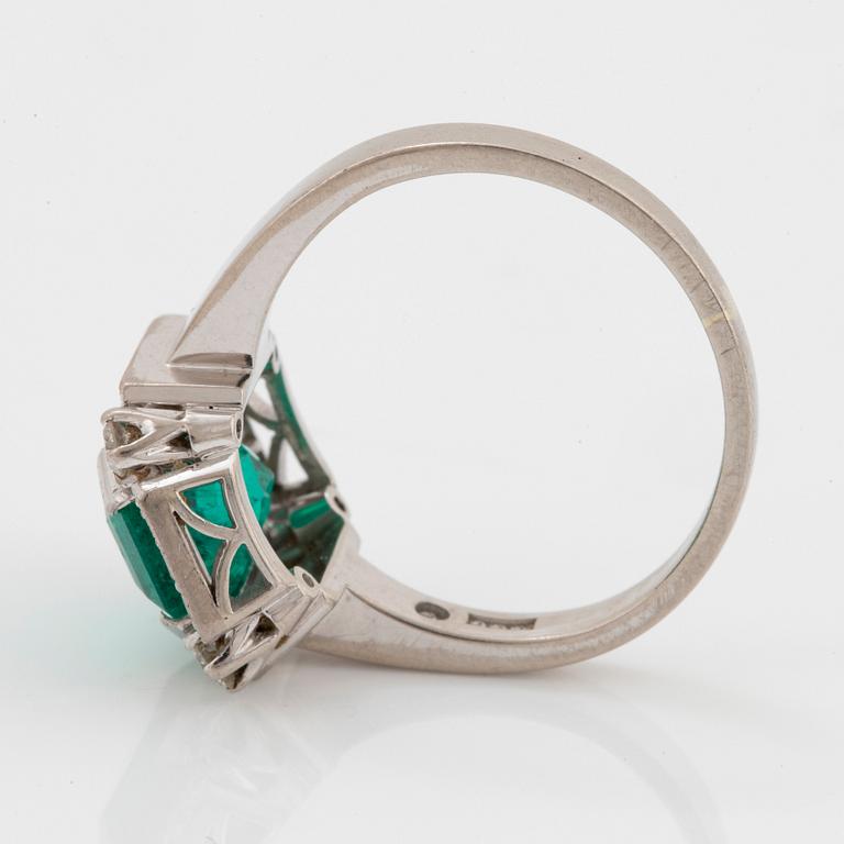 An 18K white gold ring set with a faceted emerald and round brilliant-cut diamonds.