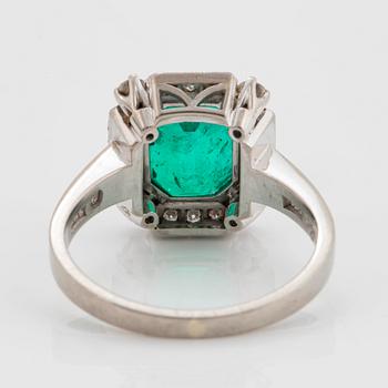 An 18K white gold ring set with a faceted emerald and round brilliant-cut diamonds.