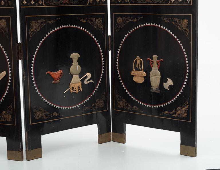 A chinese black lacquer six-panel screen, early 20th Century, with figures in gardens, inlays of carved mother of pearl, coloured bone, tree, and different stones. Back of panels with flowers painted in gold.