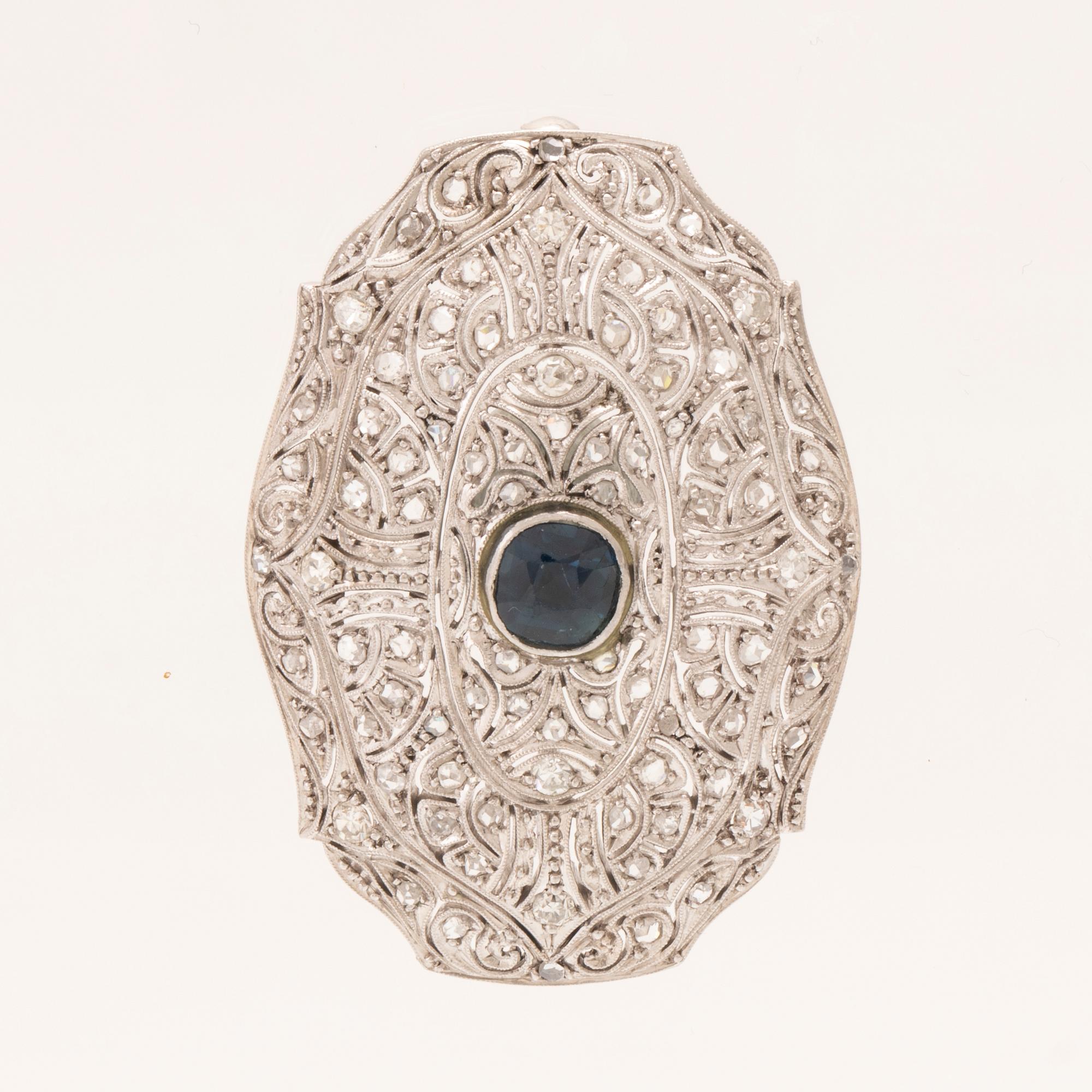 An 18K white gold brooch set with a facet-cut sapphire possibly synthetic, as well as brilliant and rose-cut diamonds.