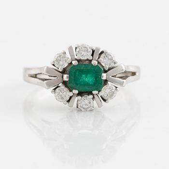 An 18K white gold ring set with a faceted emerald and round briliiant-cut diamonds.