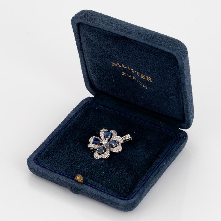 A Meister brooch in 18K white gold set with faceted sapphires, a round brilliant-cut diamond.