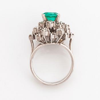 A platinum ring set with a faceted emerald and round brilliant-cut diamonds.