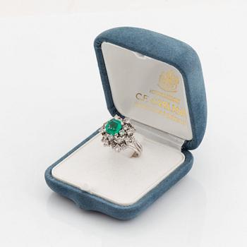 A platinum ring set with a faceted emerald and round brilliant-cut diamonds.