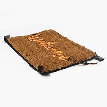 Banksy, dörrmatta, handbroderad med material från livvästar, 2019.