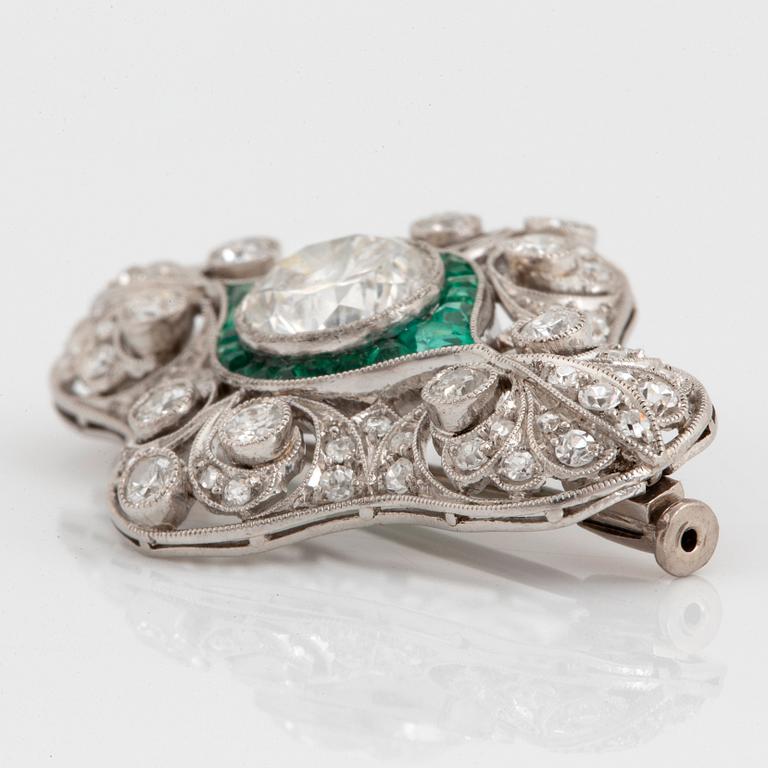 A platinum brooch set with round brilliant- and old-cut diamonds and faceted emeralds.