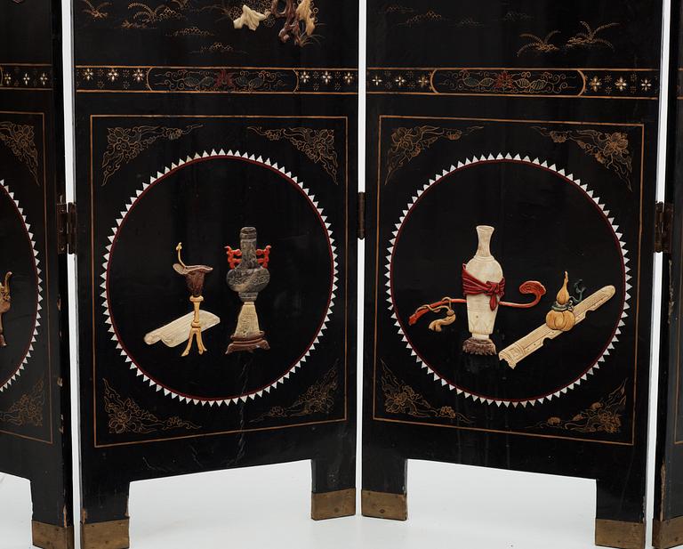 A chinese black lacquer six-panel screen, early 20th Century, with figures in gardens, inlays of carved mother of pearl, coloured bone, tree, and different stones. Back of panels with flowers painted in gold.