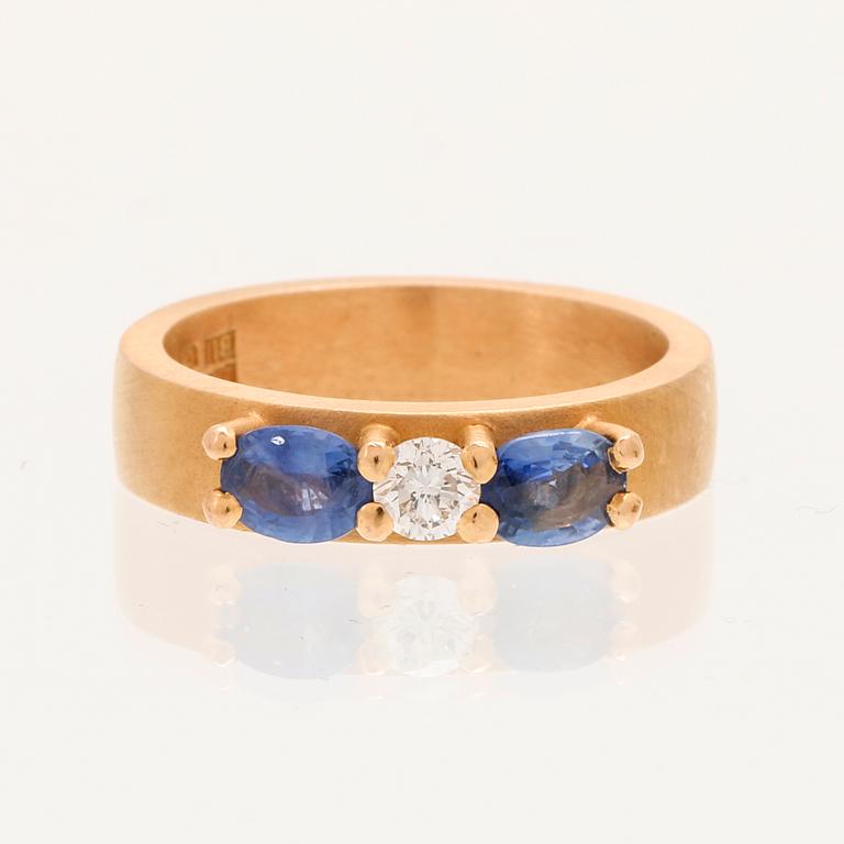 A ring containing at least 21.6K gold, set with a round cut brilliant diamond and oval brilliant cut sapphires.