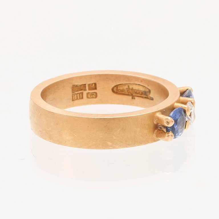 A ring containing at least 21.6K gold, set with a round cut brilliant diamond and oval brilliant cut sapphires.
