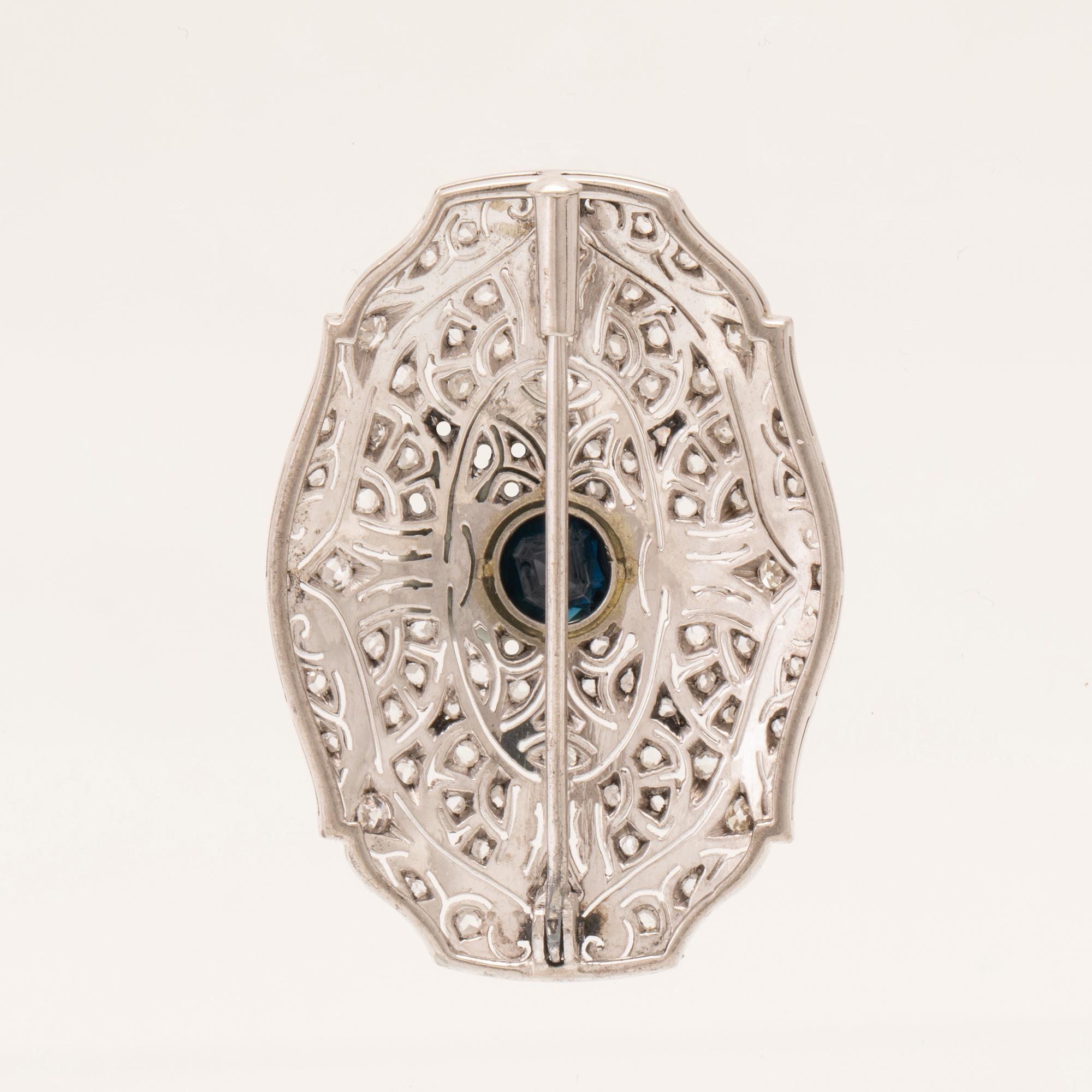 An 18K white gold brooch set with a facet-cut sapphire possibly synthetic, as well as brilliant and rose-cut diamonds.