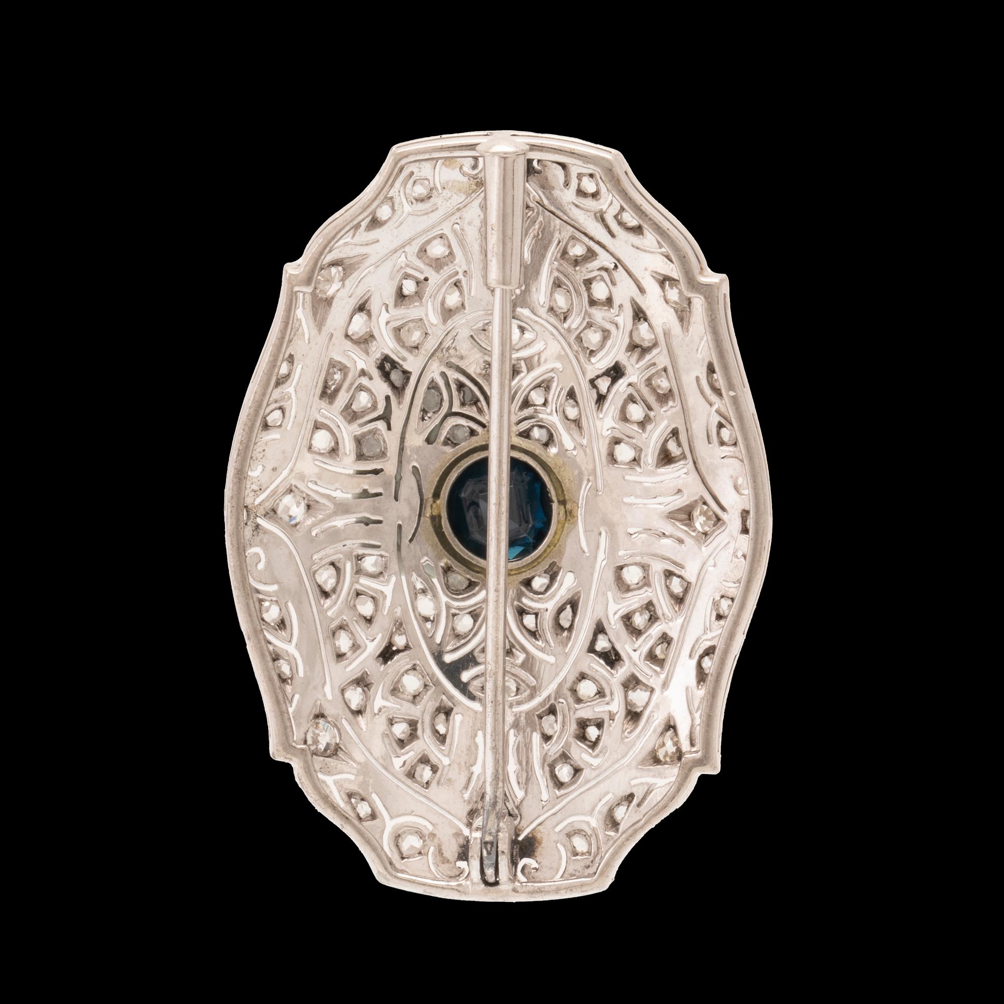 An 18K white gold brooch set with a facet-cut sapphire possibly synthetic, as well as brilliant and rose-cut diamonds.