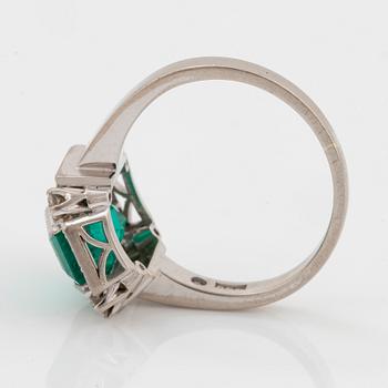 An 18K white gold ring set with a faceted emerald and round brilliant-cut diamonds.