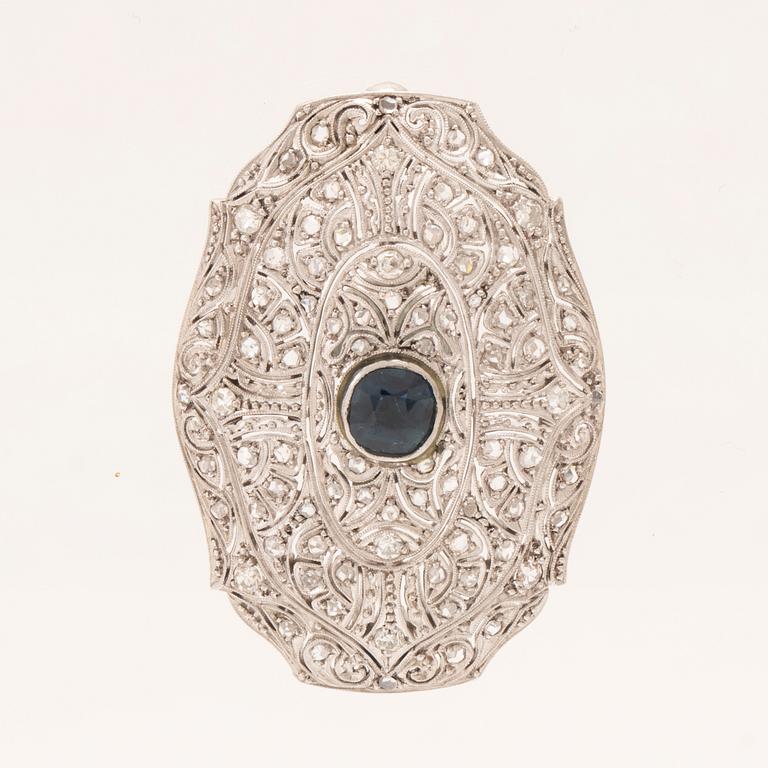 An 18K white gold brooch set with a facet-cut sapphire possibly synthetic, as well as brilliant and rose-cut diamonds.