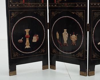 A chinese black lacquer six-panel screen, early 20th Century, with figures in gardens, inlays of carved mother of pearl, coloured bone, tree, and different stones. Back of panels with flowers painted in gold.