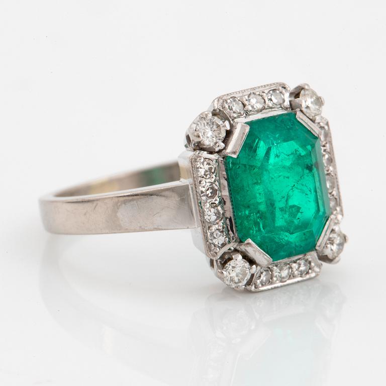 An 18K white gold ring set with a faceted emerald and round brilliant-cut diamonds.