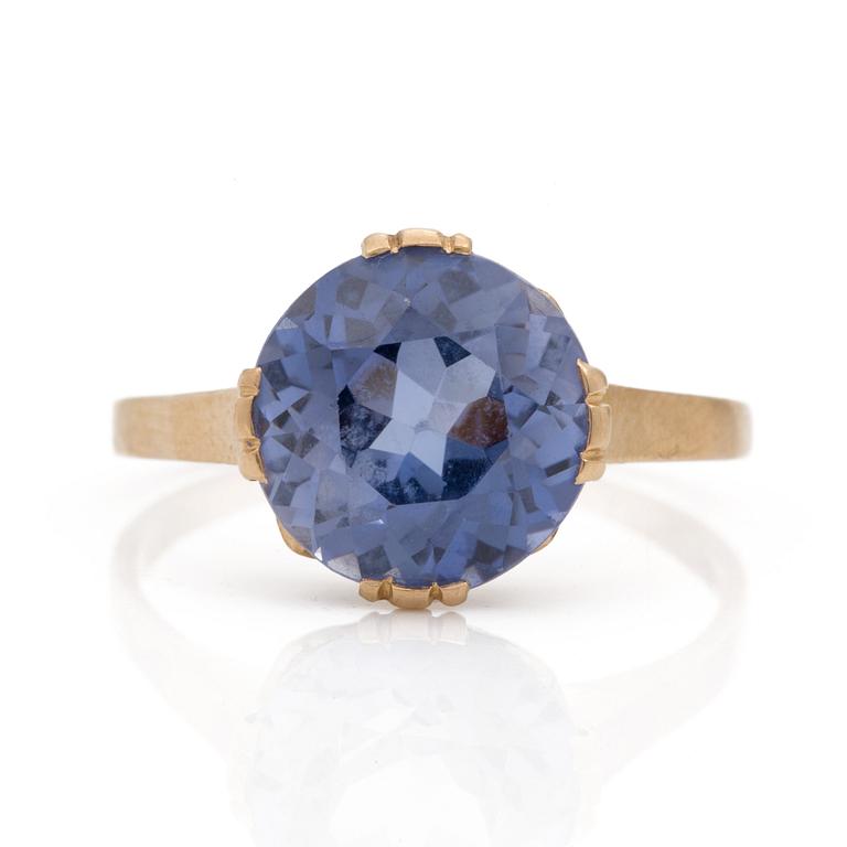 A ring set with a round, mixed-cut, synthetic sapphire.